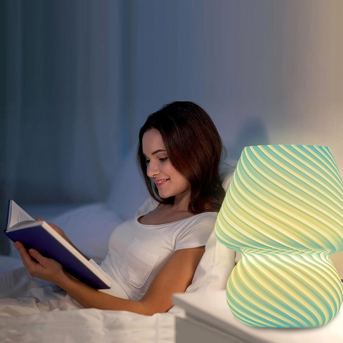Eine Frau liest im Bett neben einer beleuchteten, grün-gelb gewirbelten Lampe. Sie lächelt. Der Hintergrund ist eine dunkelblaue Wand.