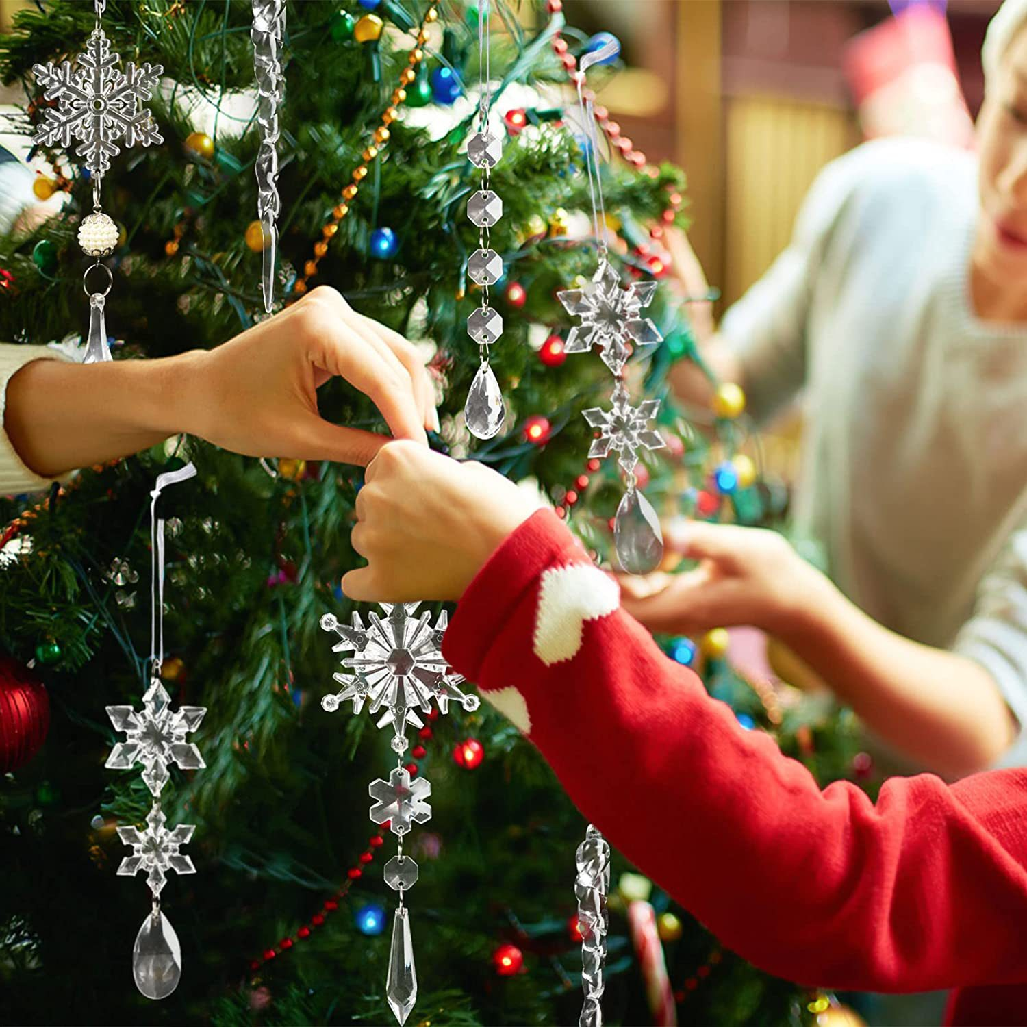 Hände schmücken einen Weihnachtsbaum mit Schneeflocken-Ornamenten. Eine Person trägt einen roten Pullover. Der Baum ist mit bunten Lichtern beleuchtet.