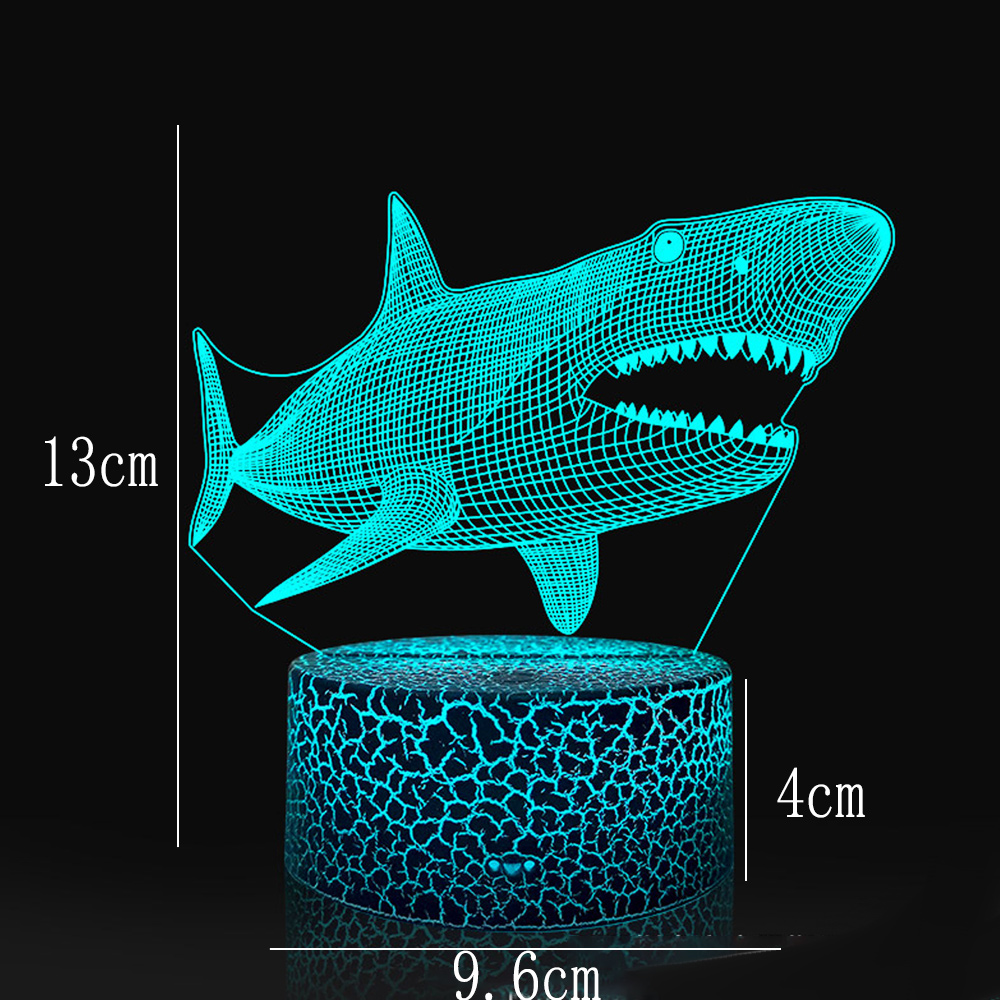 Eine Haifischlampe mit blauem Umriss. Sie steht auf einem Sockel mit Rissmuster. Die Maße sind 13 cm hoch, 9,6 cm breit, und der Sockel ist 4 cm.