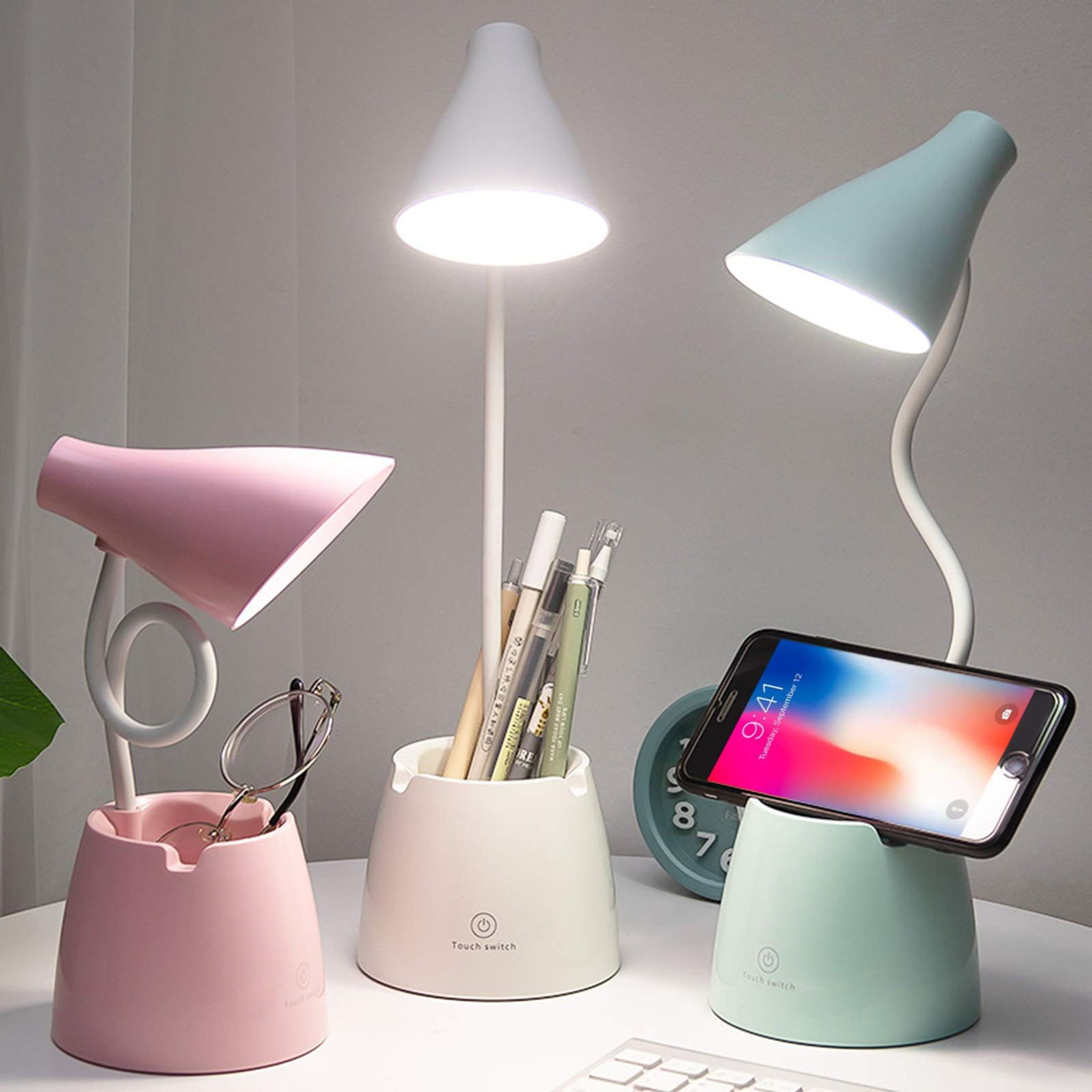 Drei Schreibtischlampen mit Stifthaltern, Brille und Telefon. Lampen sind rosa, weiß und blaugrün. Stifte im weißen Halter.