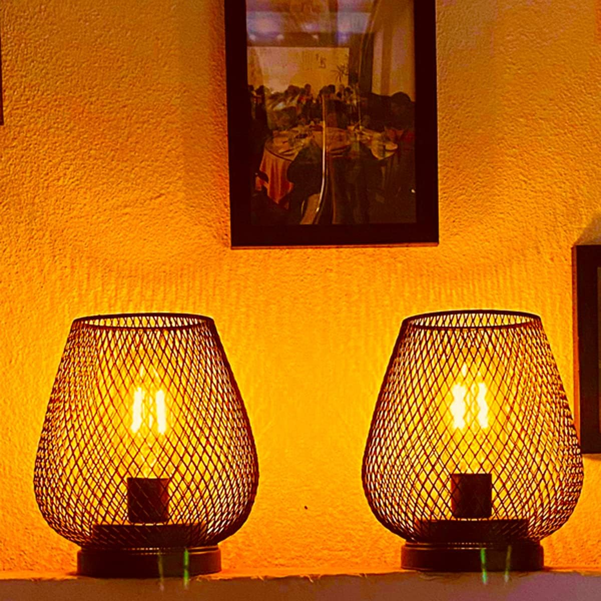 Zwei dekorative Lampen mit Gitterschirmen auf einem Regal, von innen beleuchtet. Ein gerahmtes Bild hängt darüber an einer gelben Wand.