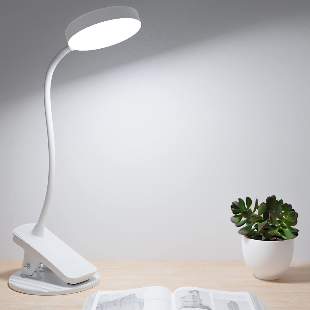 Eine weiße Schreibtischlampe mit Clipfuß steht auf einem Holztisch. Eine Pflanze in einem weißen Topf und ein offenes Buch sind ebenfalls auf dem Tisch.