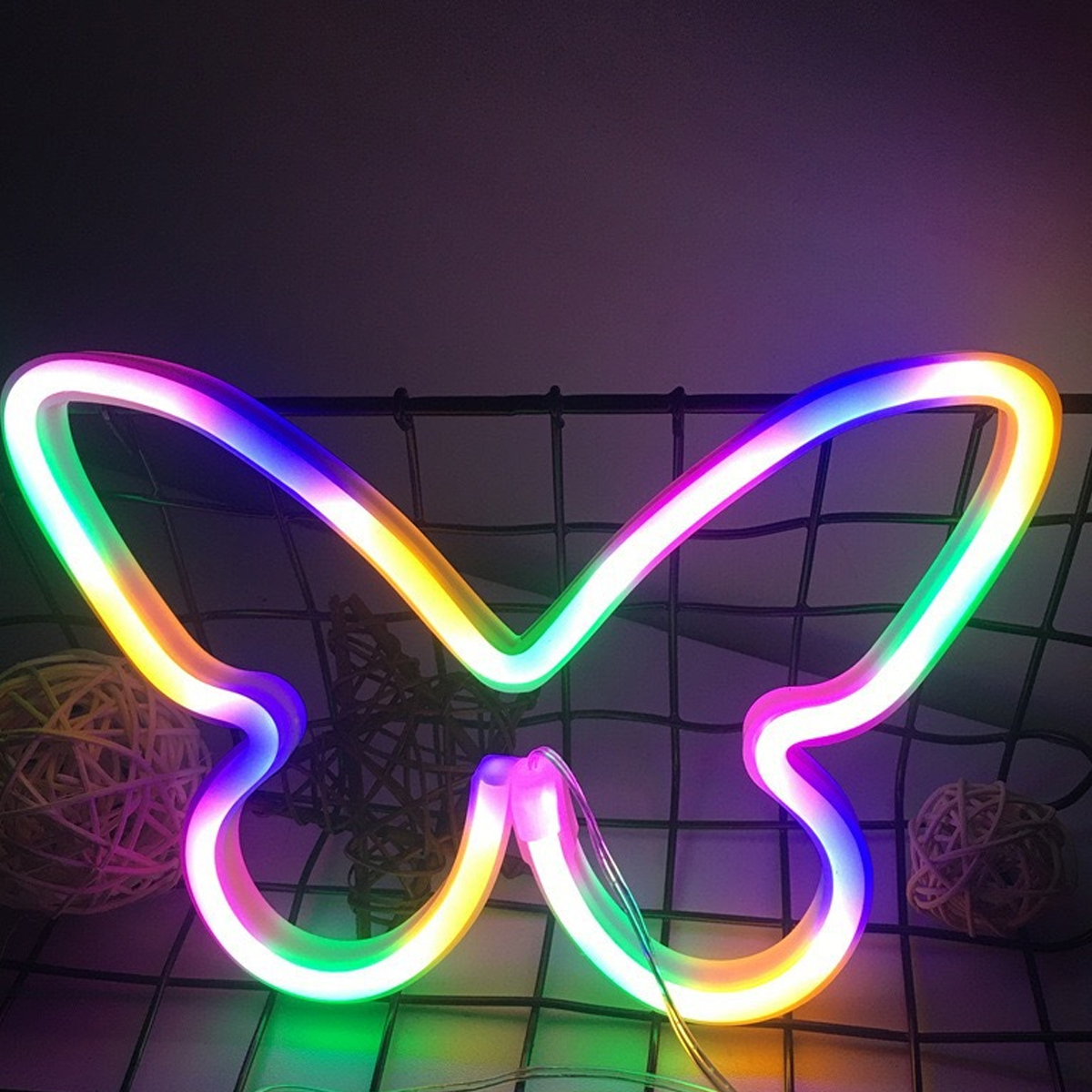 Ein Neon-Schmetterlingsschild mit Regenbogenfarben leuchtet vor dunklem Hintergrund. Korbkugeln und ein Metallgitter sind ebenfalls sichtbar.