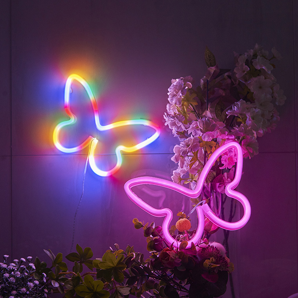 Zwei Neon-Schmetterlingslichter, eines mehrfarbig, eines rosa, neben Blumen.