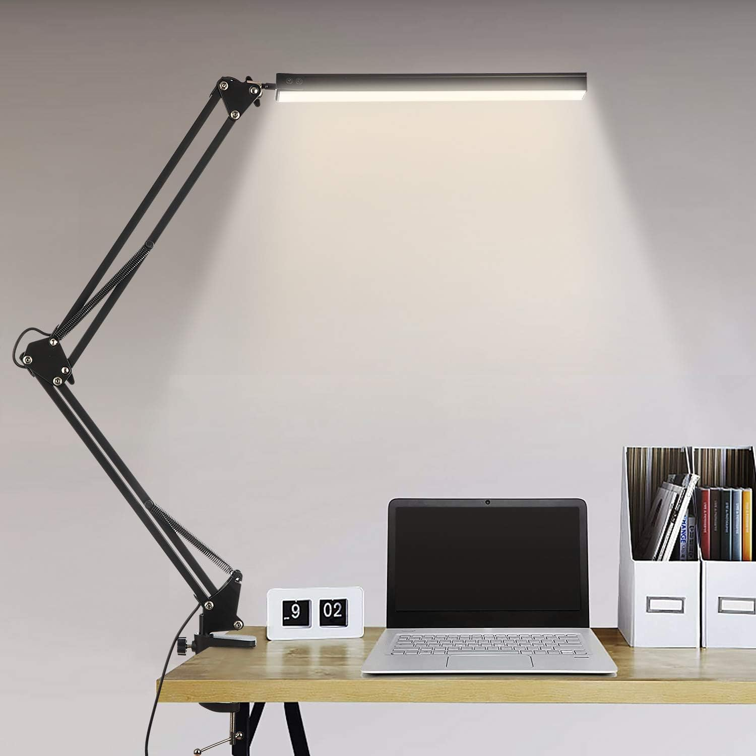 Ein Schreibtisch mit Laptop, Lampe, Uhr und Dokumentenhaltern. Die schwarze Lampe ist auf den Schreibtisch gerichtet.