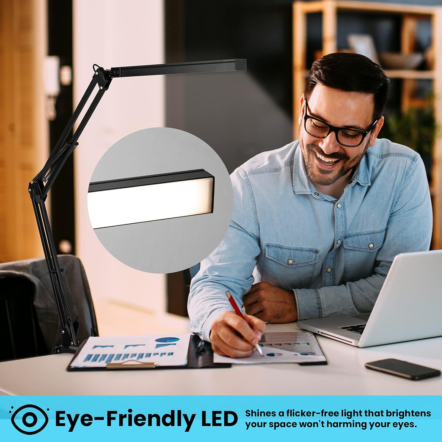 Ein Mann mit Brille lächelt und schreibt auf Papier. Eine Schreibtischlampe ist über ihm. Laptop, Telefon und Papiere sind sichtbar.