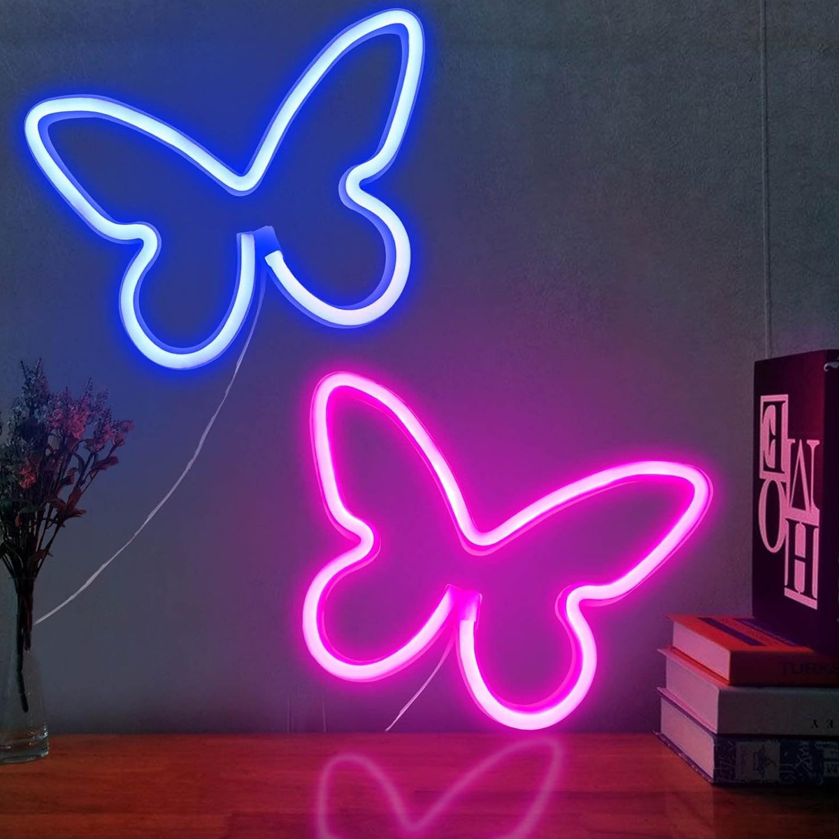 Zwei Neon-Schmetterlingslichter, eines blau, eines pink, an einer Wand. Bücher und Blumen stehen auf einer Holzoberfläche.