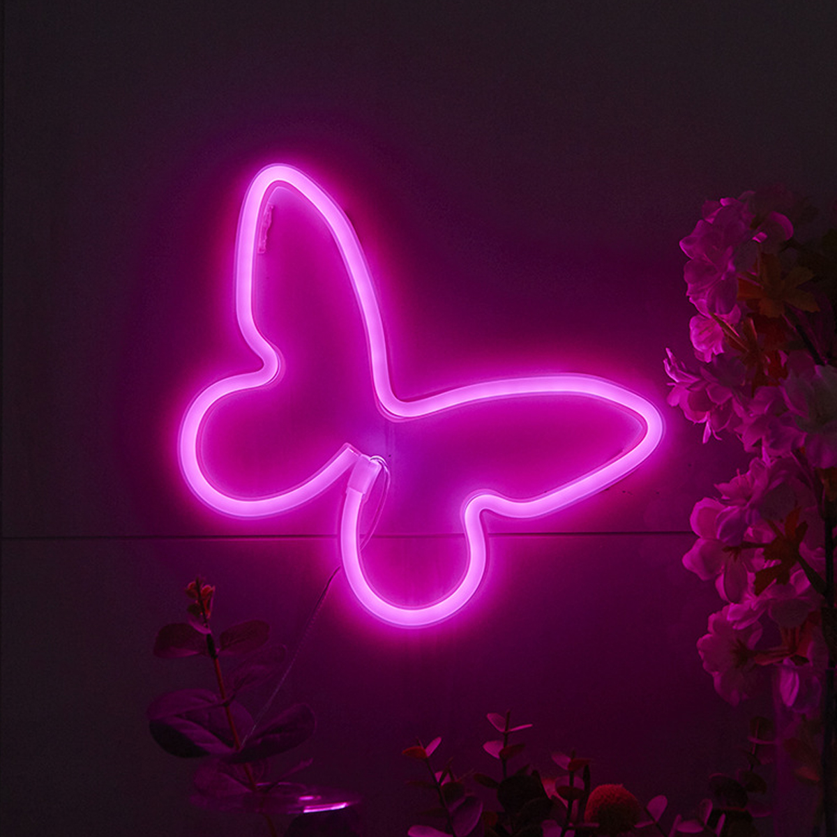Eine neonrosa Schmetterlingslichtdekoration verziert eine dunkle Wand. Rosa Blumen und grüne Blätter sind im Hintergrund.
