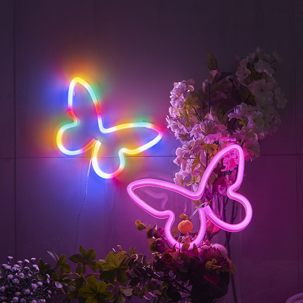 Zwei Neon-Schmetterlingslichter in leuchtenden Farben, mit Blumen im Hintergrund. Eines ist regenbogenfarben, das andere rosa.