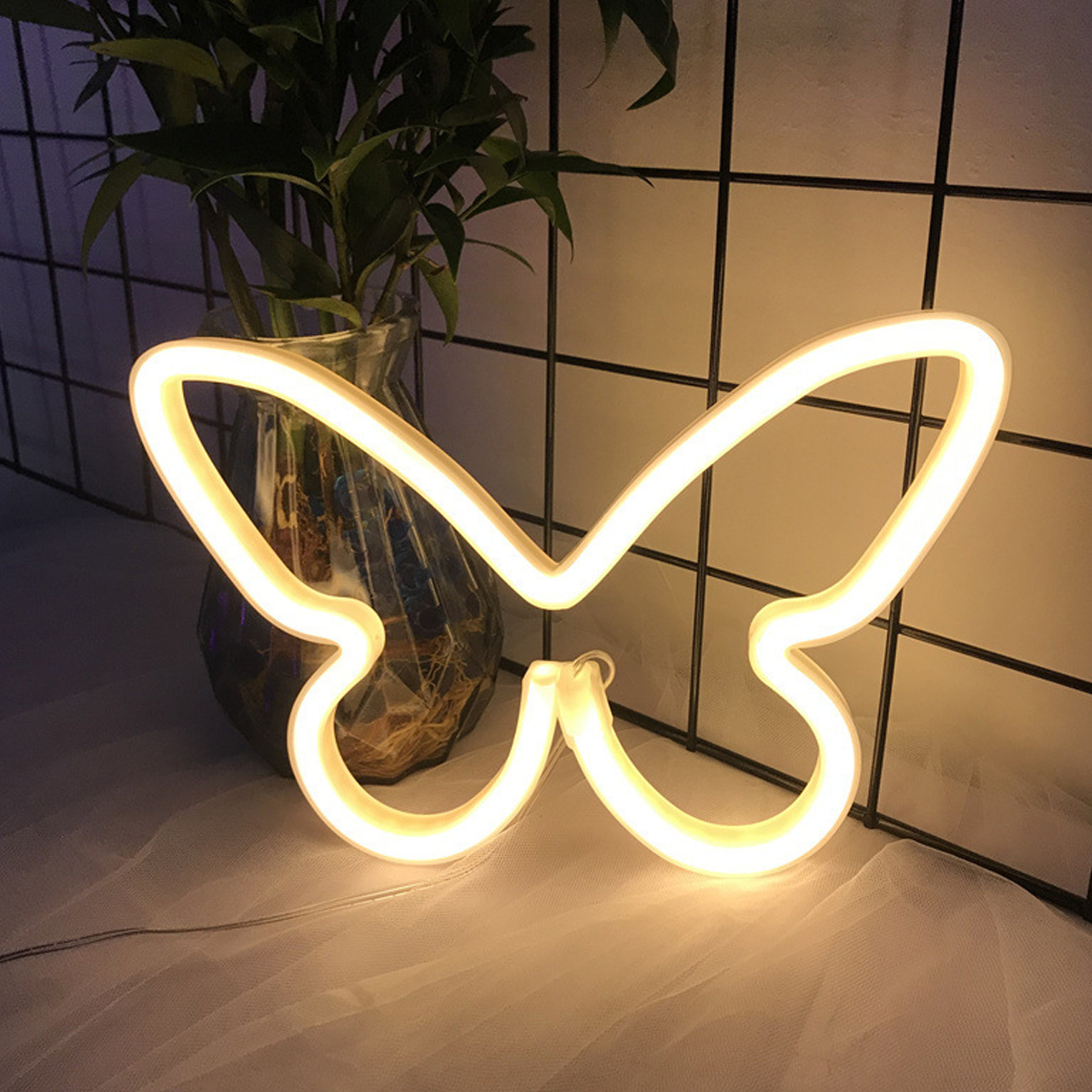 Eine leuchtende Neon-Schmetterlingsleuchte steht auf einem Tisch. Dahinter eine Vase mit Pflanzen vor einer gefliesten Wand.
