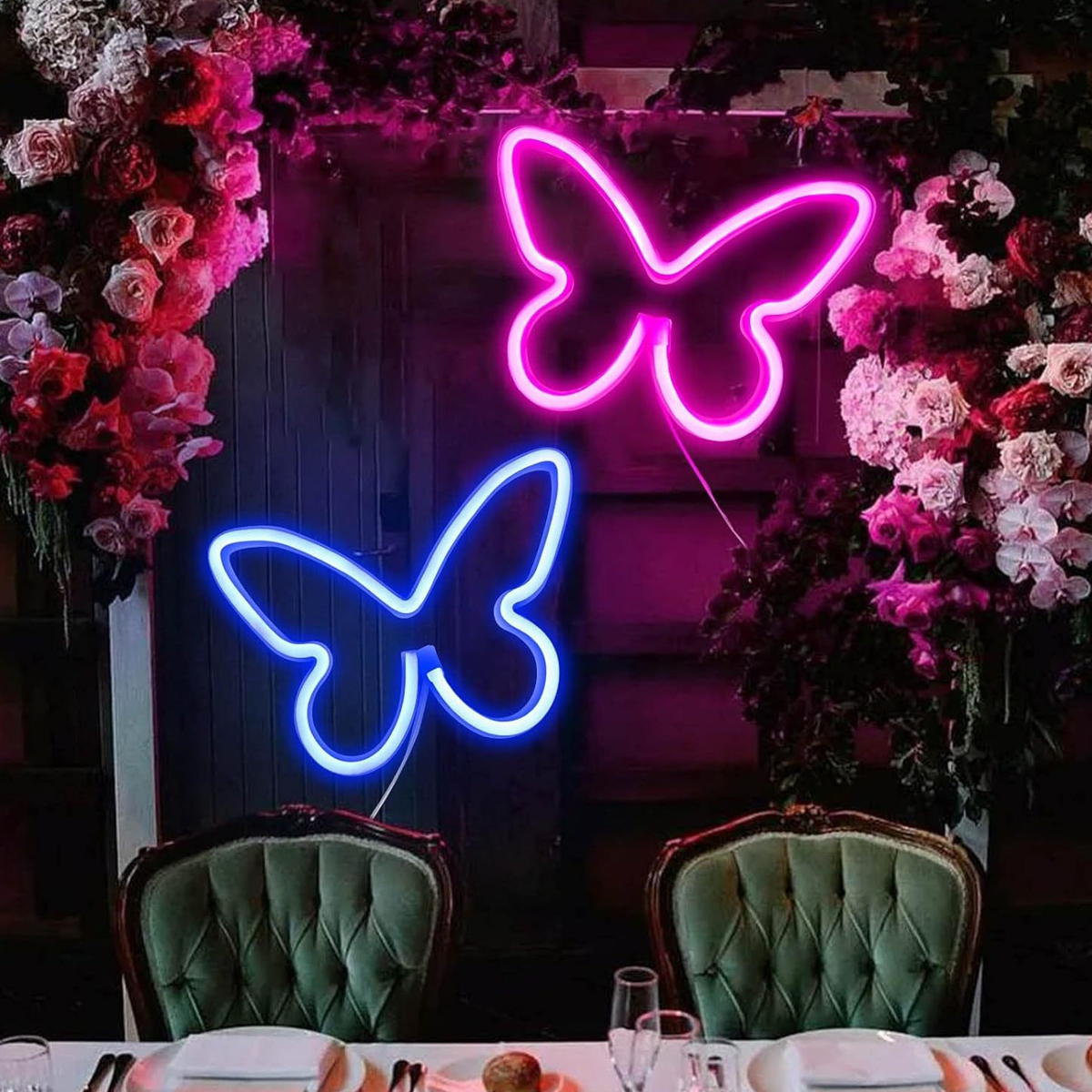 Zwei Neon-Schmetterlingslichter, rosa und blau, mit Blumen und Stühlen im Vordergrund.