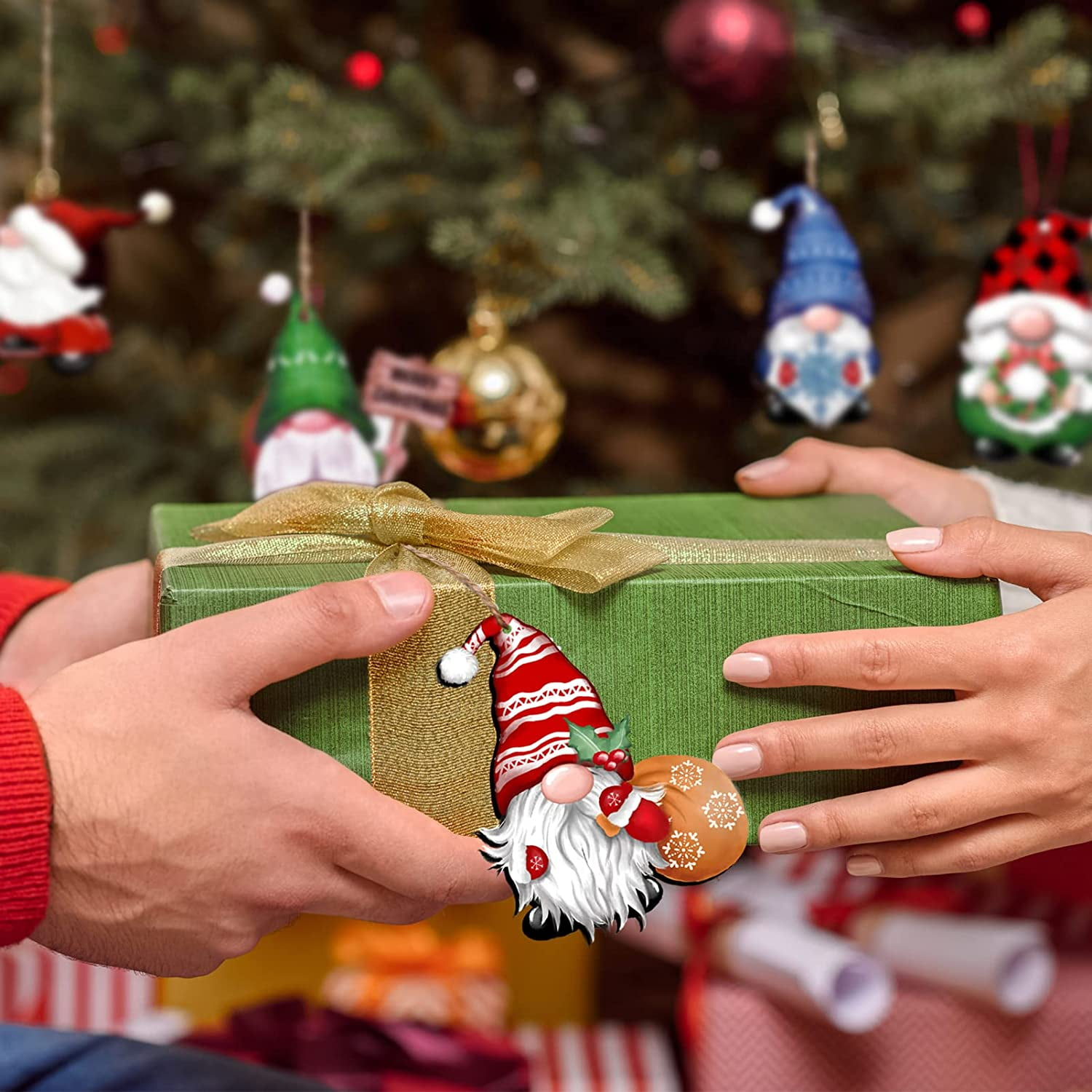 Hände tauschen eine grüne Geschenkbox mit goldener Schleife aus, Weihnachtsbaum im Hintergrund mit Ornamenten und Wichteln.