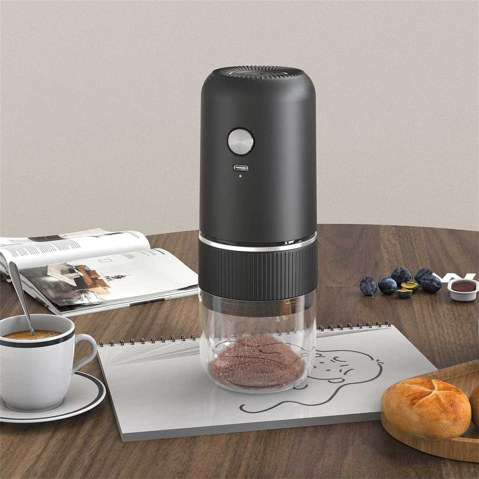 Eine Kaffeemühle steht auf einem Holztisch. Eine Tasse Kaffee, Pflaumen und ein Brötchen sind in der Nähe.