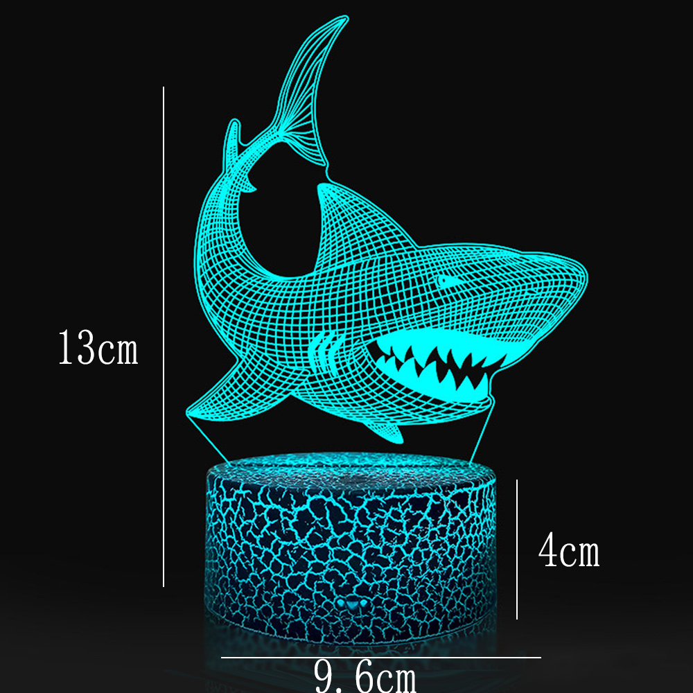Eine 3D-LED-Lampe in Form eines Haifischs. Sie leuchtet blau, Abmessungen sind angegeben. Der Hai steht auf einem rissig aussehenden Sockel.