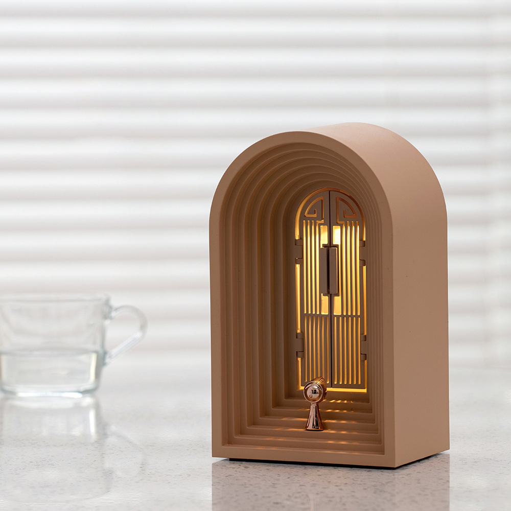 Eine beige bogenförmige Lampe mit goldenem Türdesign. Links ein Glas Wasser. Hintergrund ist ein weißer Rollladen.