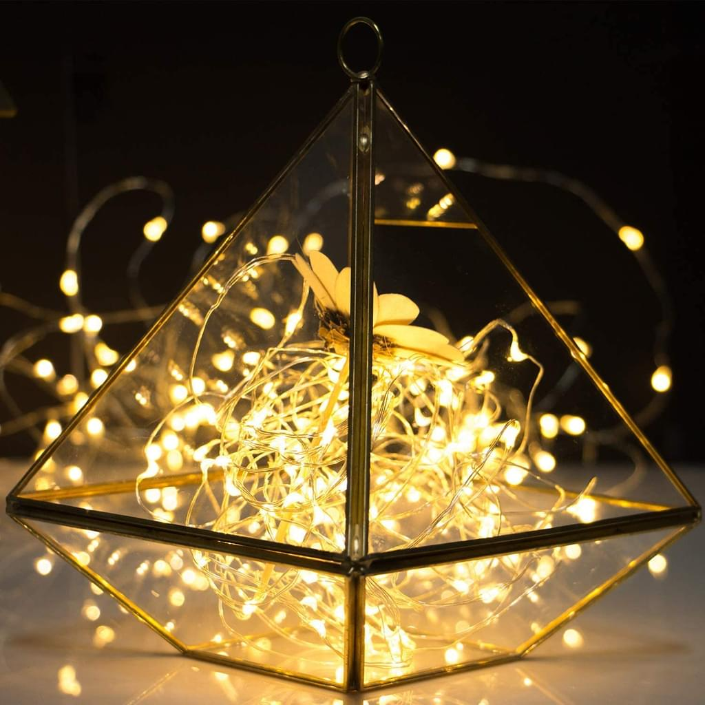 Ein dreieckiges Glas-Terrarium enthält Lichterketten und eine dekorative Blume, die vor einem dunklen Hintergrund beleuchtet werden.