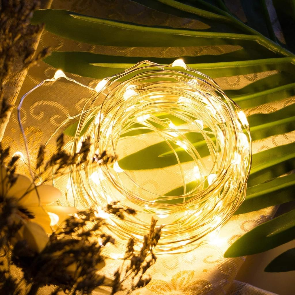 Eine Spule mit warmweißen Lichterketten liegt auf einem gemusterten Stoff, umgeben von grünen Blättern und getrockneten Blumen.