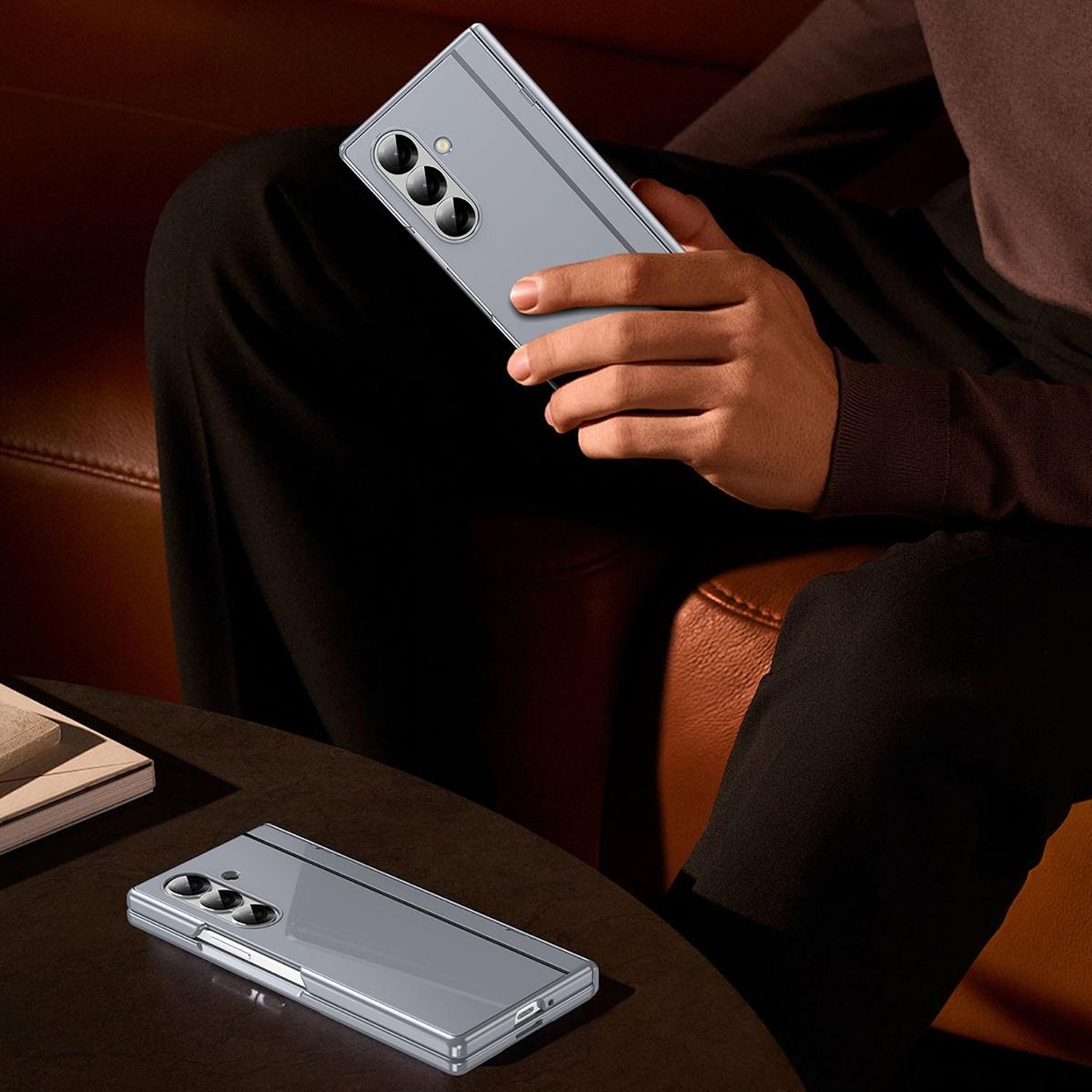 Una persona sostiene un teléfono gris plegable. El teléfono está en una mesa. La persona lleva un suéter marrón y pantalones negros.