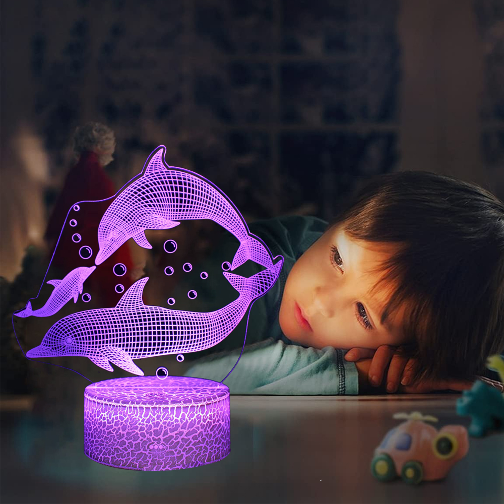 Ein Kind betrachtet eine 3D-Lampe in Delfinform. Die Lampe leuchtet lila mit Blasen. Spielzeug liegt auf dem Tisch verstreut.
