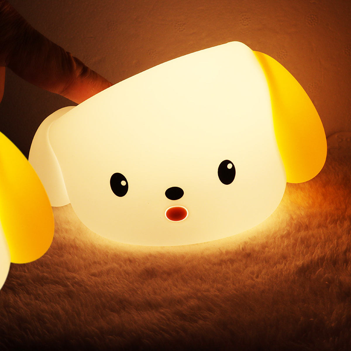 Eine leuchtende hundeförmige Lampe mit schwarzen Augen, rotem Mund und gelben Ohren liegt auf einer flauschigen Oberfläche. Eine Hand ist teilweise sichtbar.