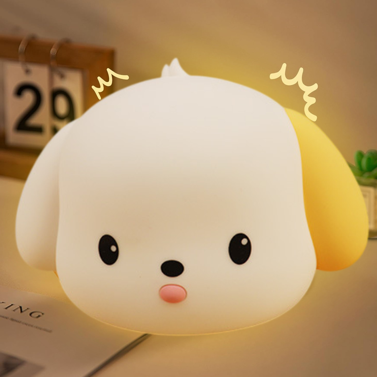 Ein weißes, hundeförmiges Nachtlicht mit gelbem Ohr. Das Licht ist an, mit einem Kalender und einer kleinen Pflanze im Hintergrund.