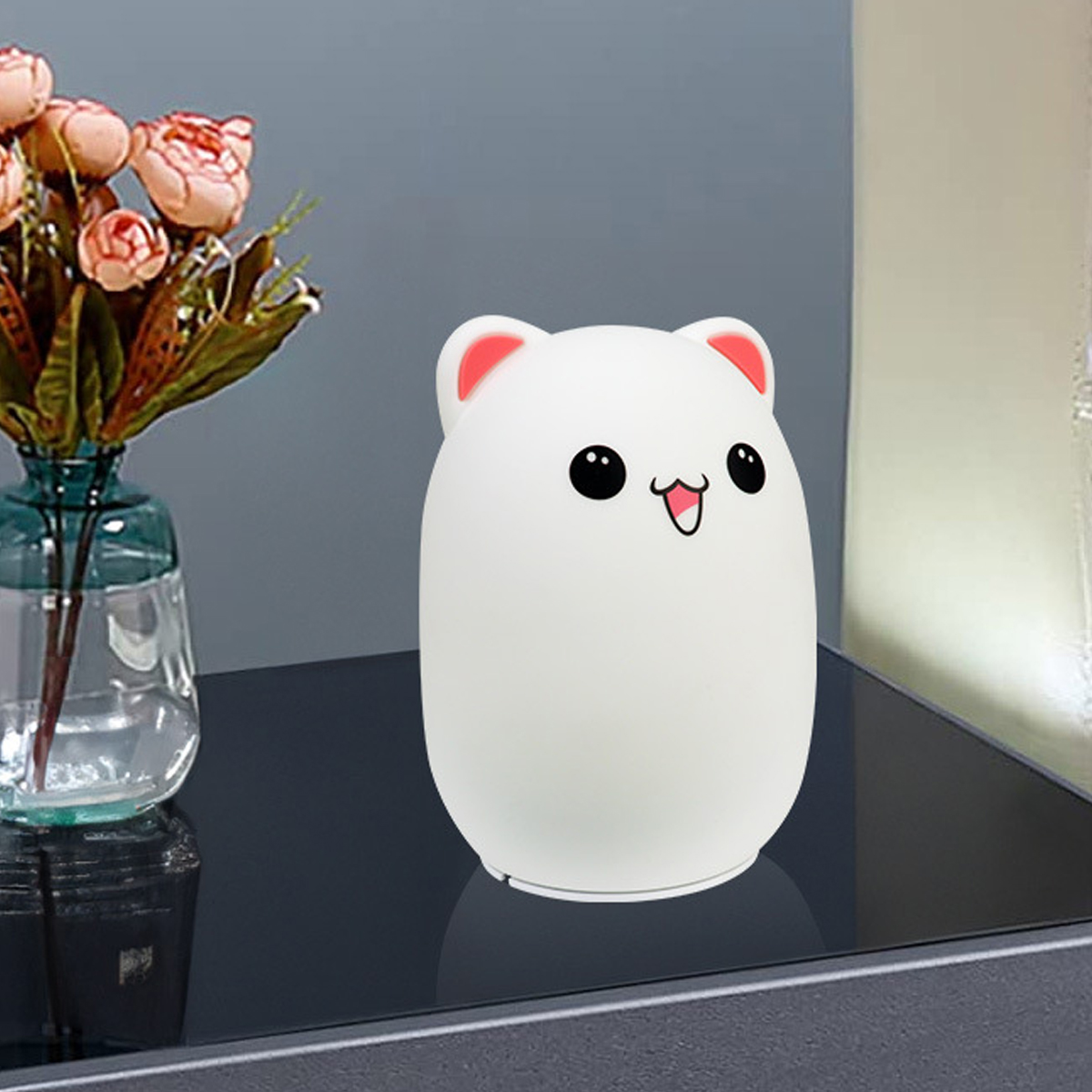 Eine weiße bärenförmige Lampe mit rosa Ohren und einem lächelnden Gesicht. Blumen in einer Vase sind links. Der Hintergrund ist eine graue Wand.