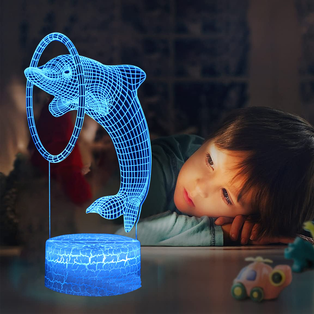 Ein Kind betrachtet eine blaue 3D-Delfinlampe. Die Lampe zeigt einen Delfin, der durch einen Reifen springt. Spielzeug liegt auf dem Tisch verstreut.