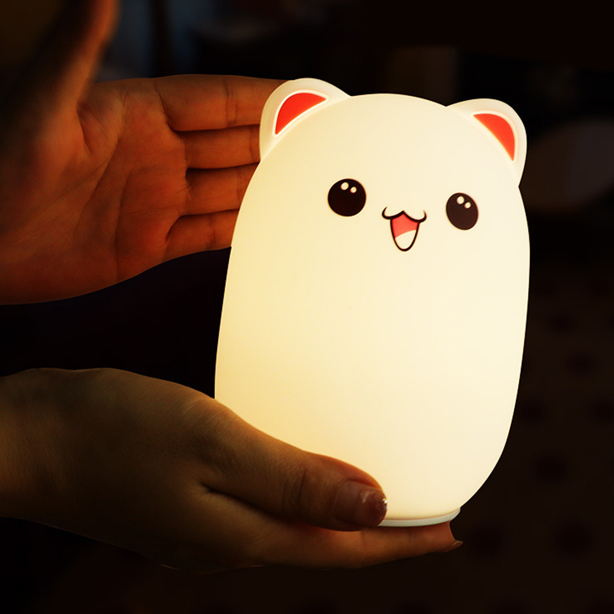 Eine Person hält eine weiße, bärenförmige Lampe mit roten Ohren. Die Lampe hat ein lächelndes Gesicht und ist beleuchtet. Der Hintergrund ist dunkel.