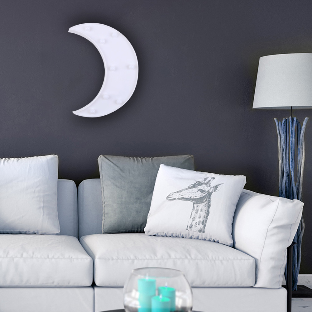 Ein weißes mondförmiges Licht hängt an einer dunklen Wand über einem weißen Sofa mit Kissen. Eine Lampe und Kerzen sind ebenfalls sichtbar.