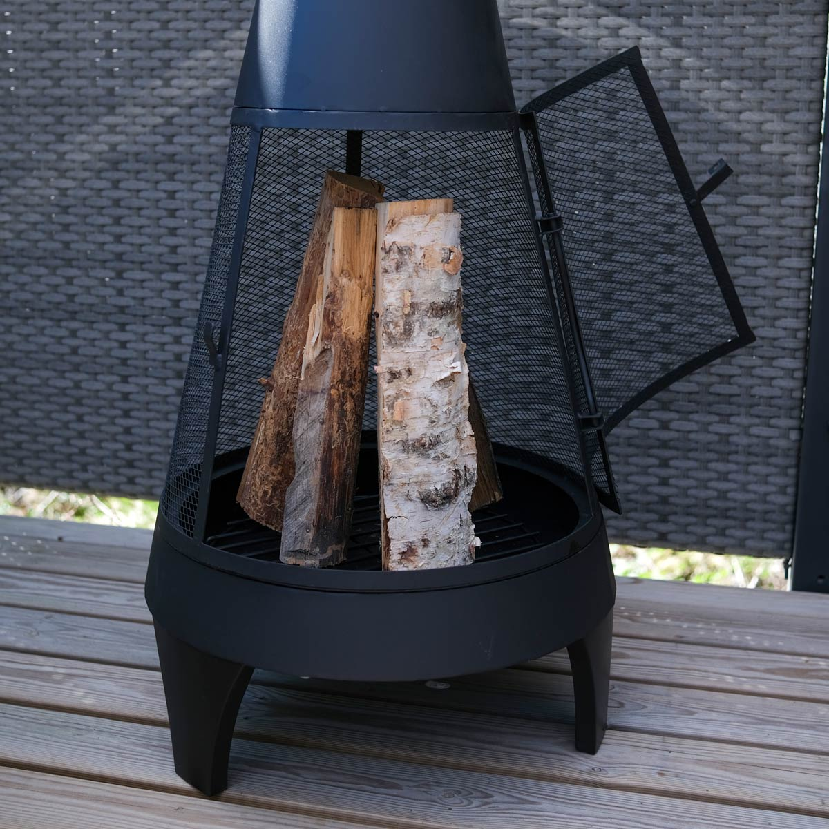 Eine schwarze Metall-Feuerstelle mit Holzscheiten im Inneren, auf einem Holzdeck. Die Tür ist offen.