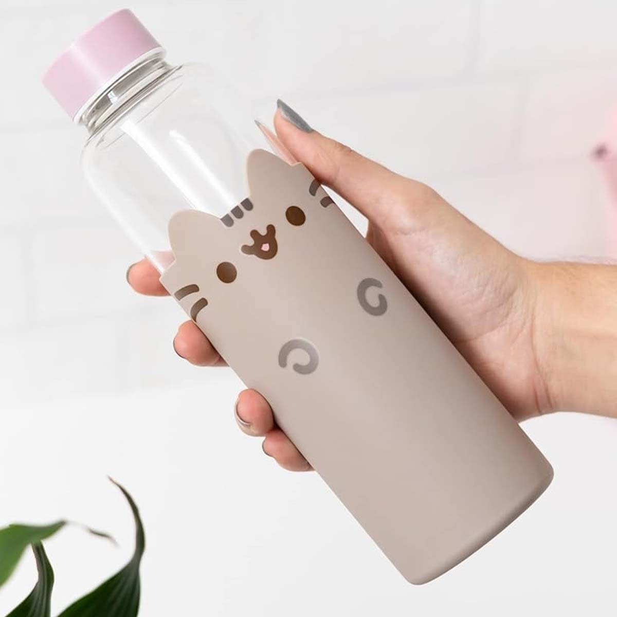 Una mano tiene in mano una bottiglia d'acqua con un design a forma di gatto. La bottiglia è trasparente con un tappo rosa e una custodia beige.