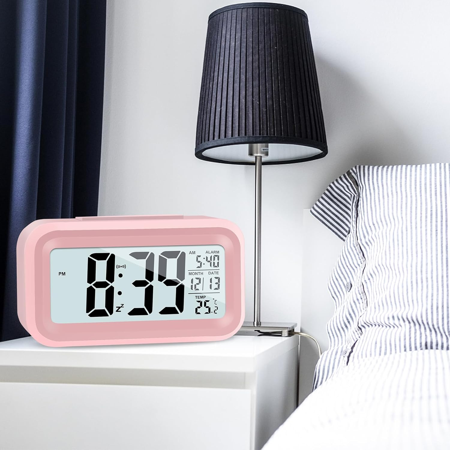 Un orologio digitale rosa mostra le 20:39. Una lampada nera è dietro su un comodino bianco. Un letto con biancheria da letto a righe è accanto.