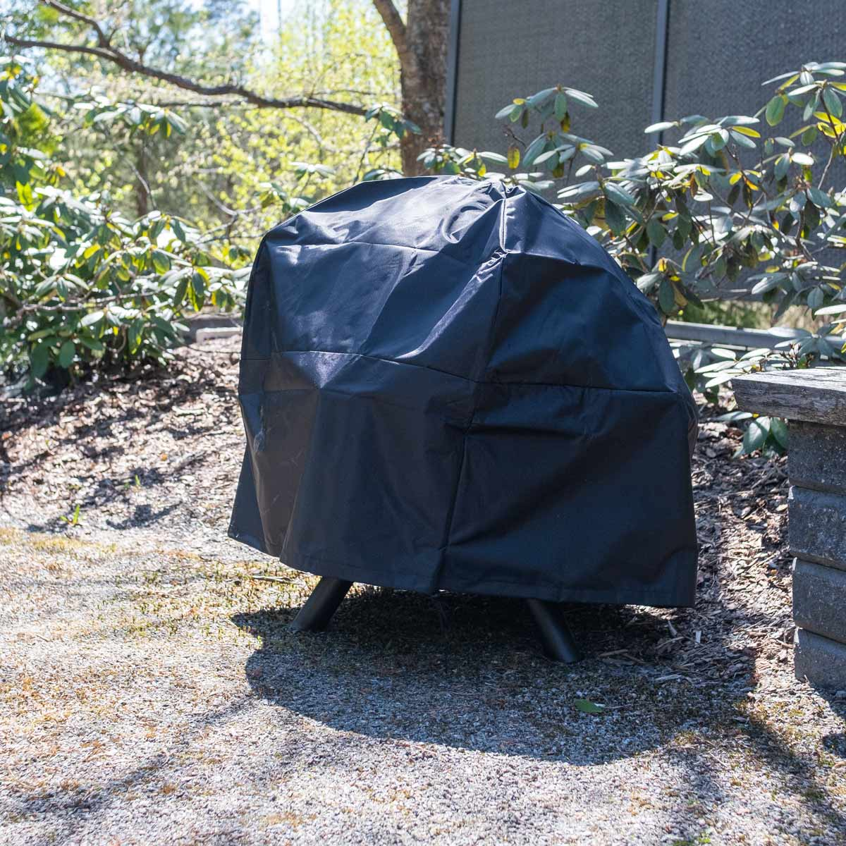 Eine schwarze Grillabdeckung steht auf einem Kiesweg in einem Garten. Bäume und Büsche umgeben sie. Rechts ist eine Holzbank.