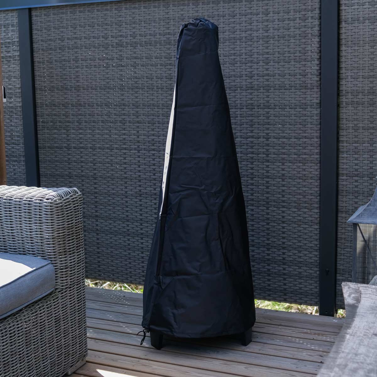 Eine hohe, schwarze Terrassenheizungsabdeckung steht auf einer Holzterrasse. Ein Korbstuhl und eine graue Wand sind ebenfalls sichtbar.