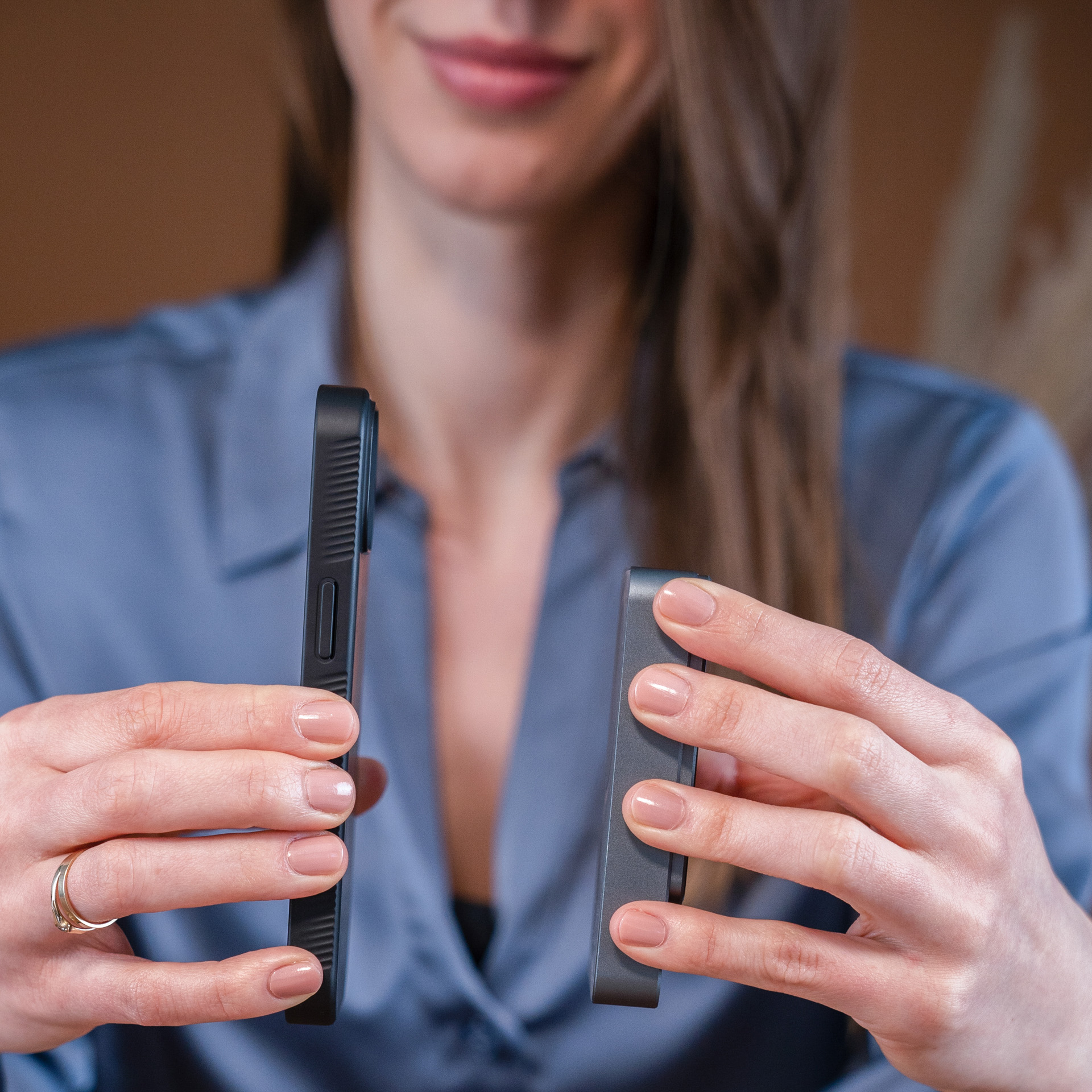 Una donna tiene in mano un telefono nero e un dispositivo rettangolare nero. Indossa una camicia blu, ha la pelle chiara e lunghi capelli castani.