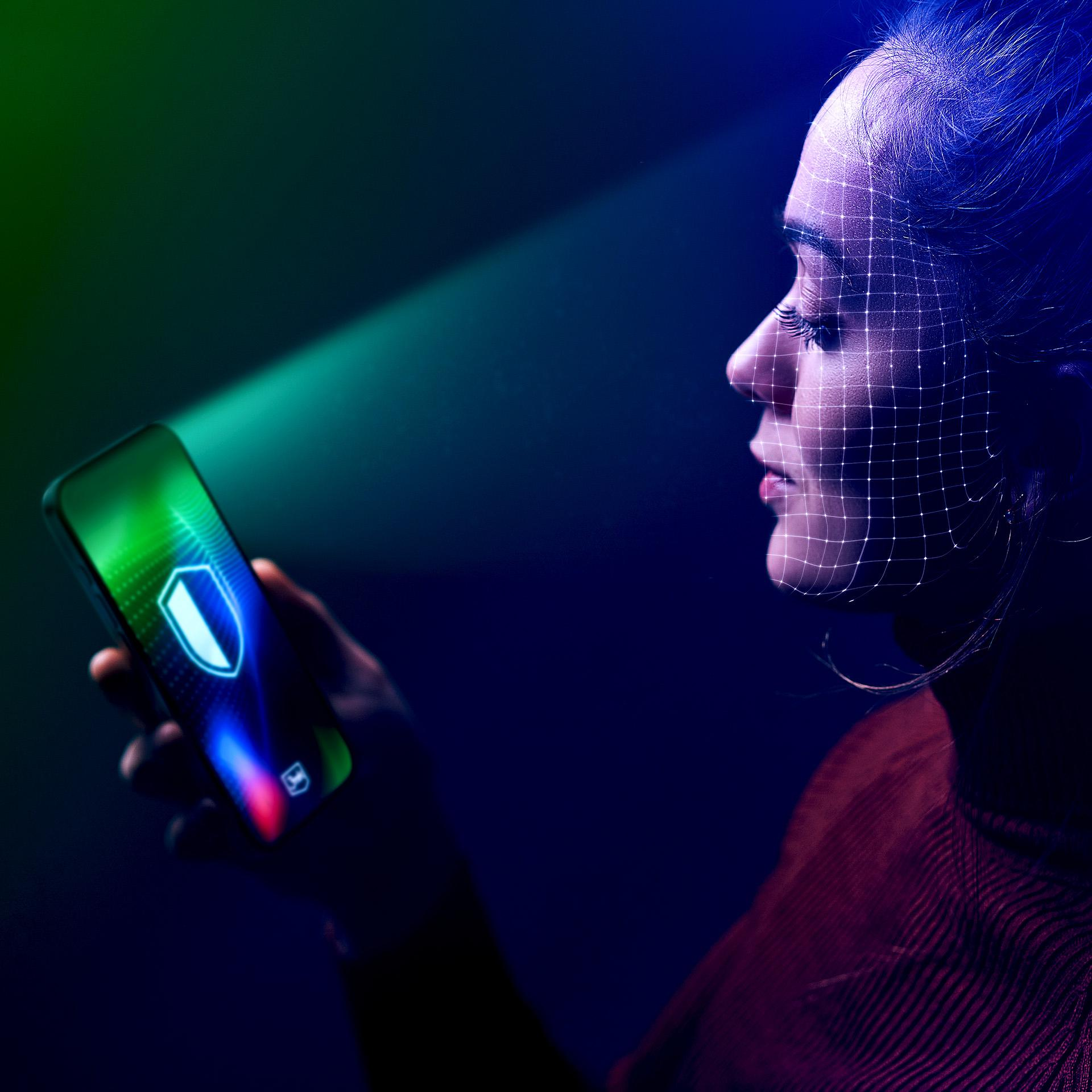 Kobieta z siatką na twarzy trzymająca telefon z ikoną tarczy, oświetlona zielonym światłem, ciemne tło.