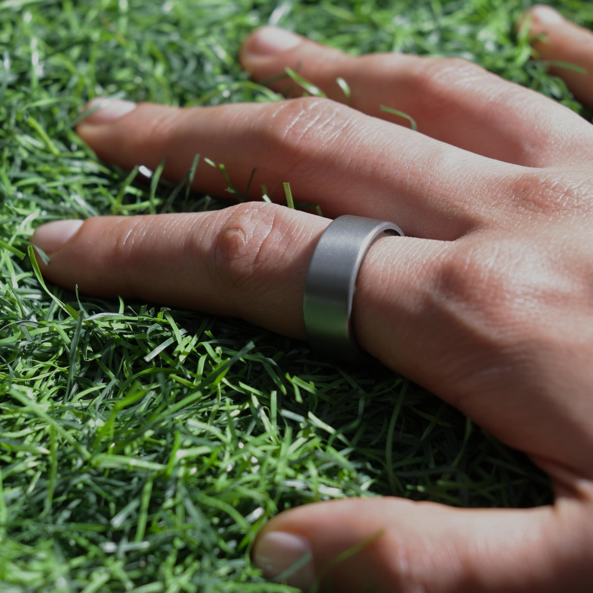 Eine Hand mit einem silbernen Ring auf einem Finger ruht auf grünem Kunstrasen. Der Ring ist ein schlichtes Band.