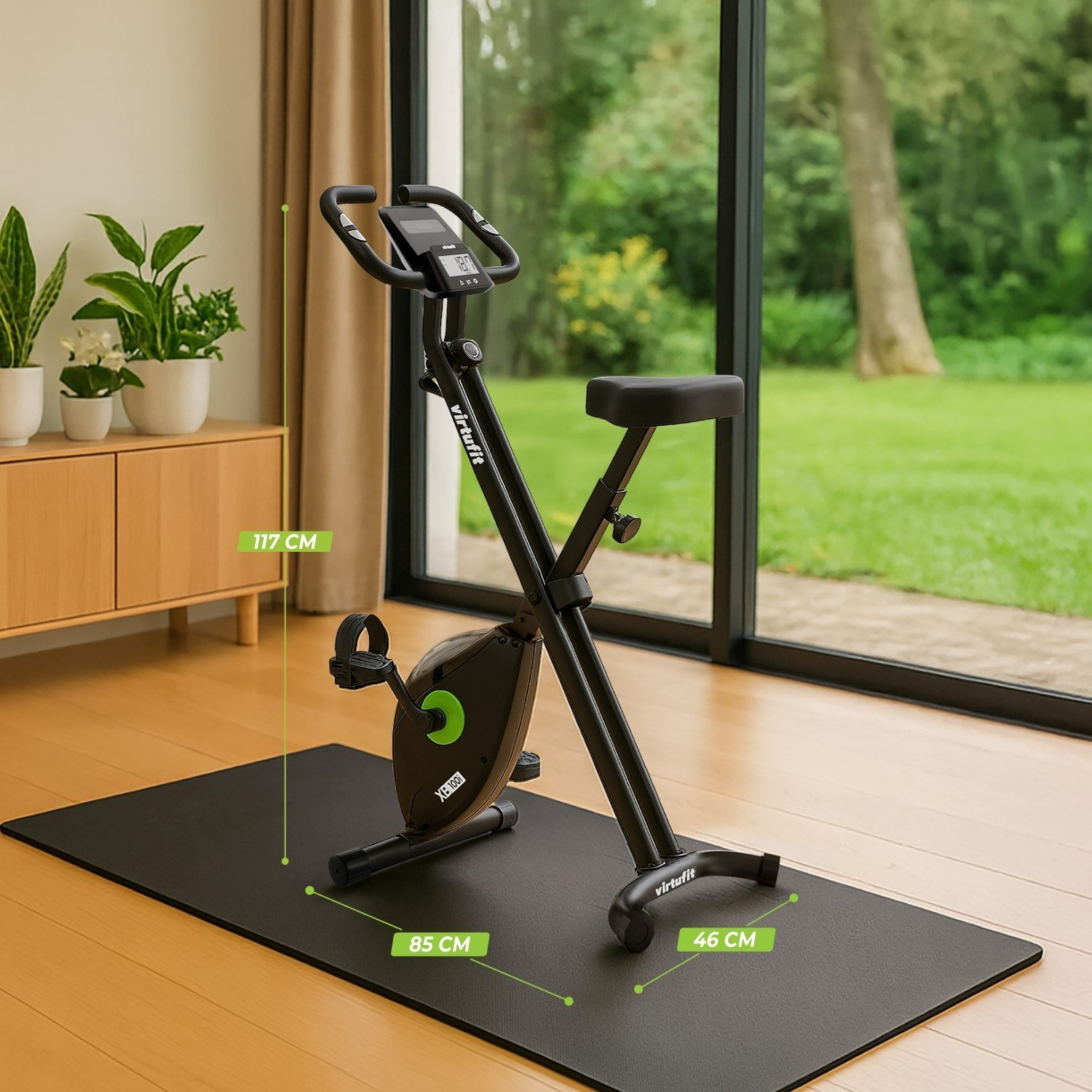 Schwarzes Trainingsfahrrad auf einer schwarzen Matte, in der Nähe eines Fensters mit Blick auf eine grüne Wiese. Pflanzen und ein Holzschrank links.