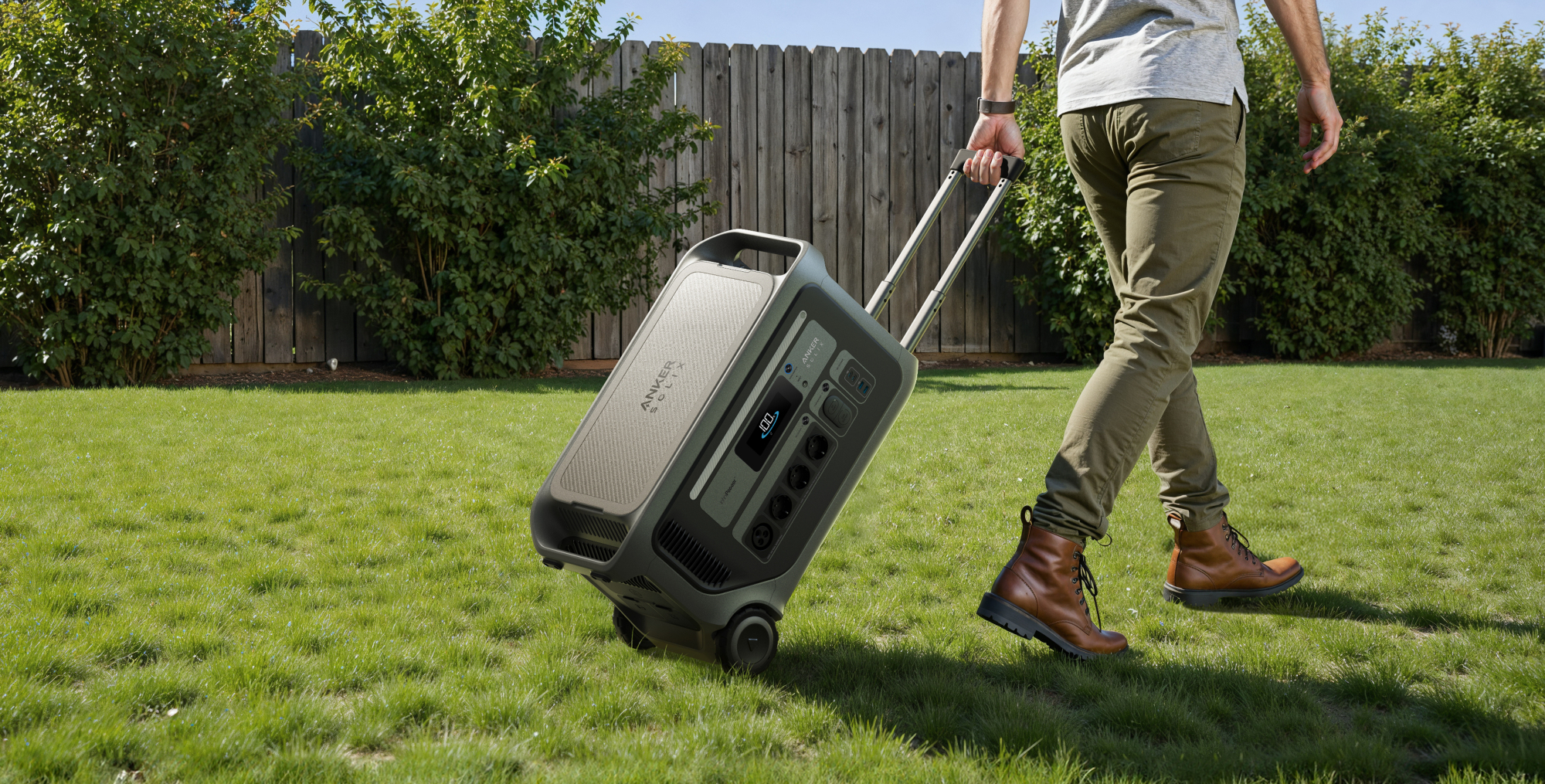 Mężczyzna ciągnie przenośną stację zasilania po trawniku. Za nim drewniane ogrodzenie i krzewy. Niebo jest niebieskie.
