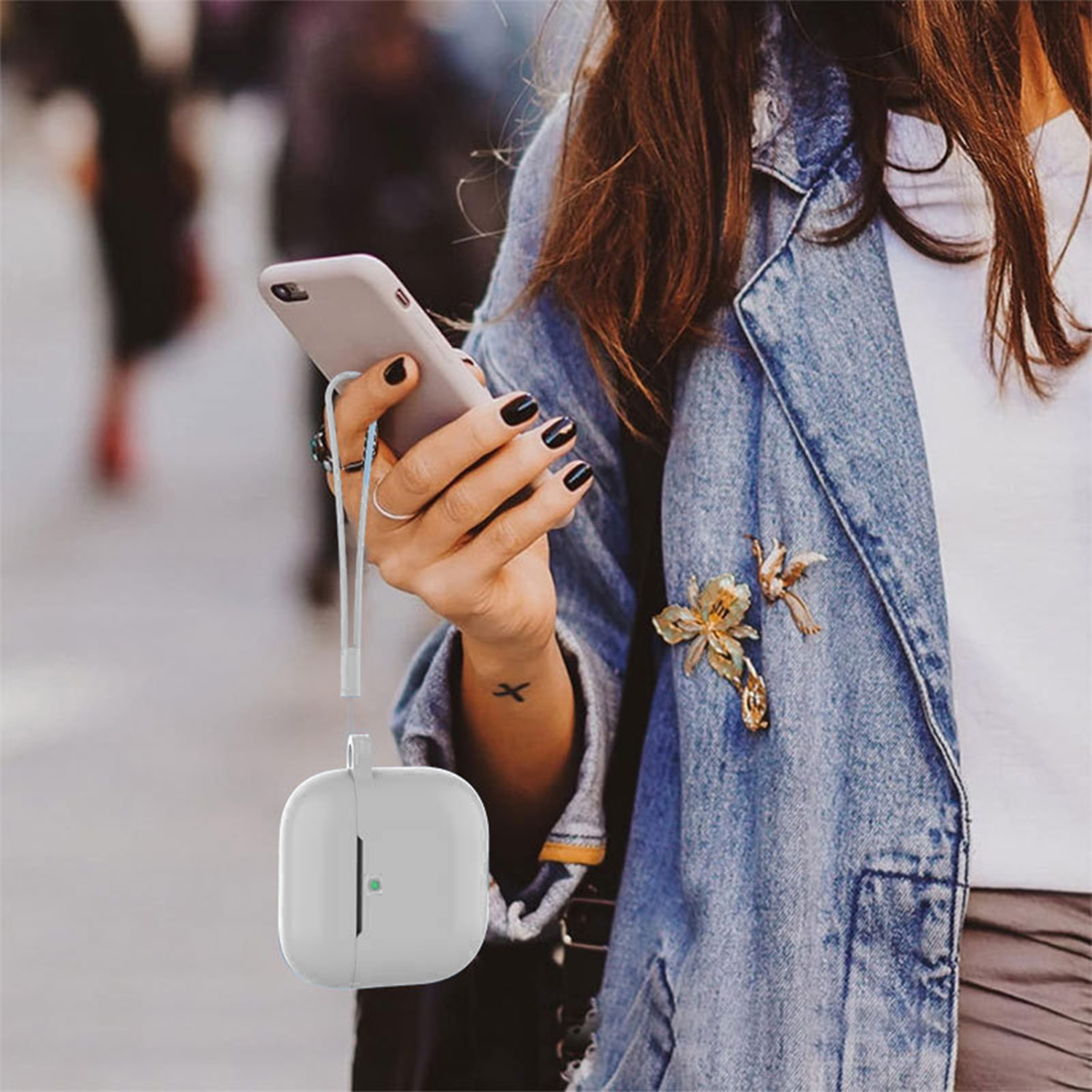 Una donna tiene in mano un telefono con auricolari. Una custodia bianca è appesa sotto. Indossa una giacca di jeans e ha un tatuaggio. Lo sfondo è sfocato.