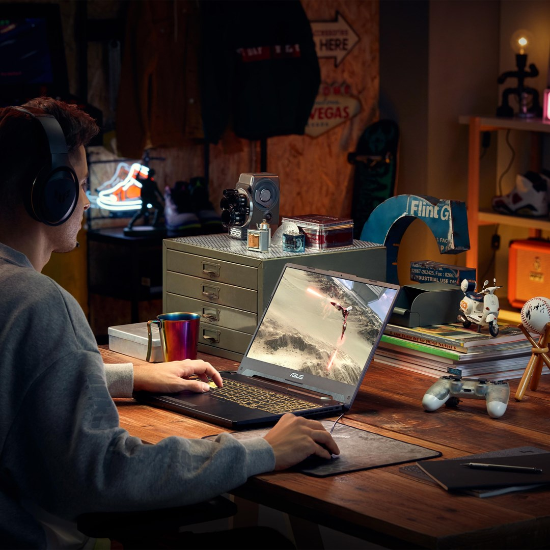 Mężczyzna w słuchawkach gra na laptopie. Pokój jest pełen dekoracji i przedmiotów kolekcjonerskich. Ekran wyświetla grę wideo.