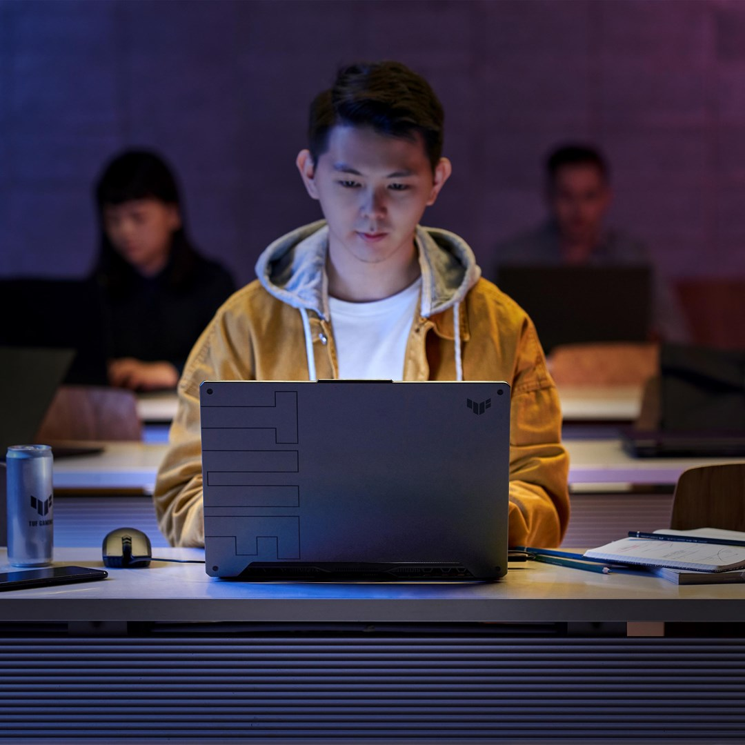 Mężczyzna w żółtej kurtce używa laptopa. Inni ludzie z laptopami są w tle. Puszka, mysz i notatnik leżą na stole.