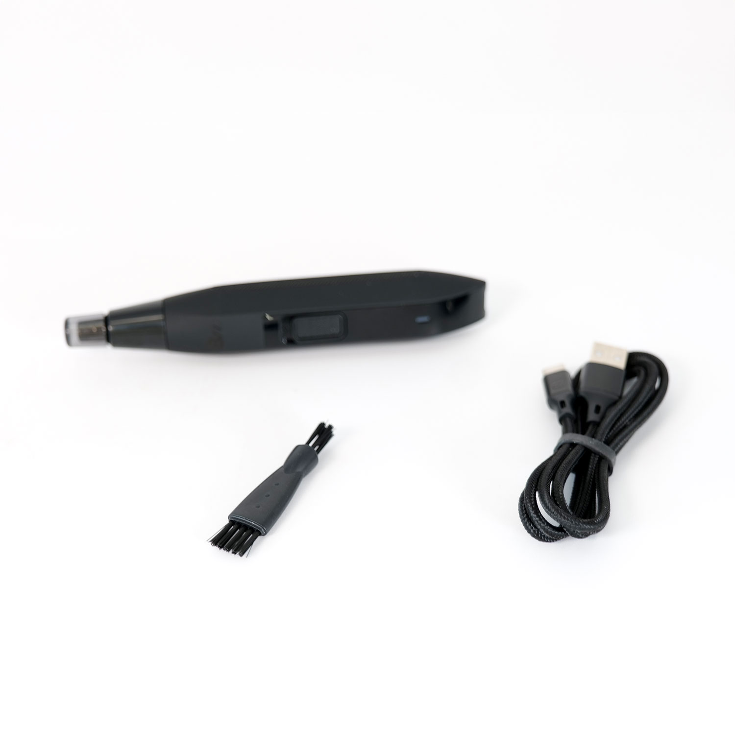 Un recortador de vello nasal negro, un cepillo pequeño y un cable USB están sobre una superficie blanca. El recortador está al fondo.