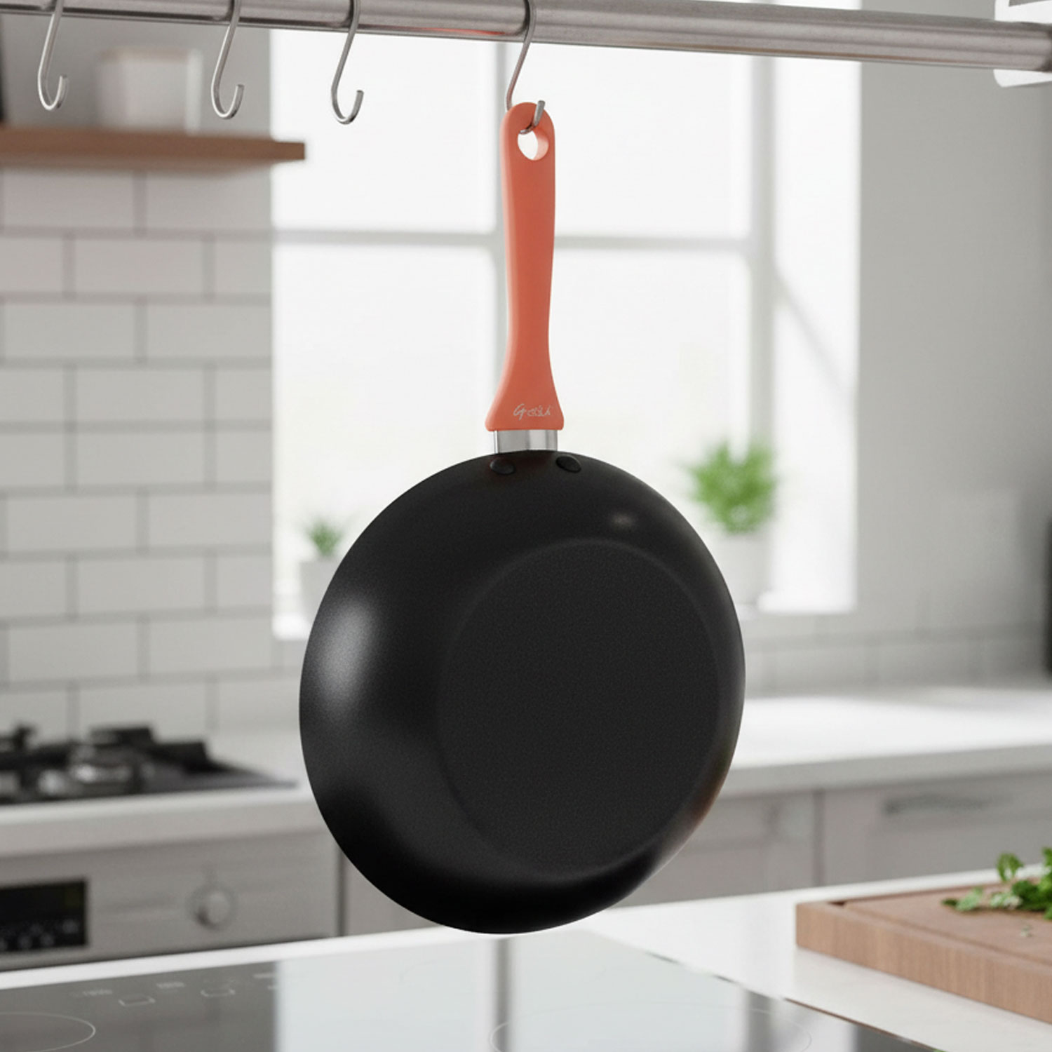 Una sartén negra con mango naranja cuelga de una barra de metal en una cocina. El fondo incluye una ventana, estufa y mostrador.