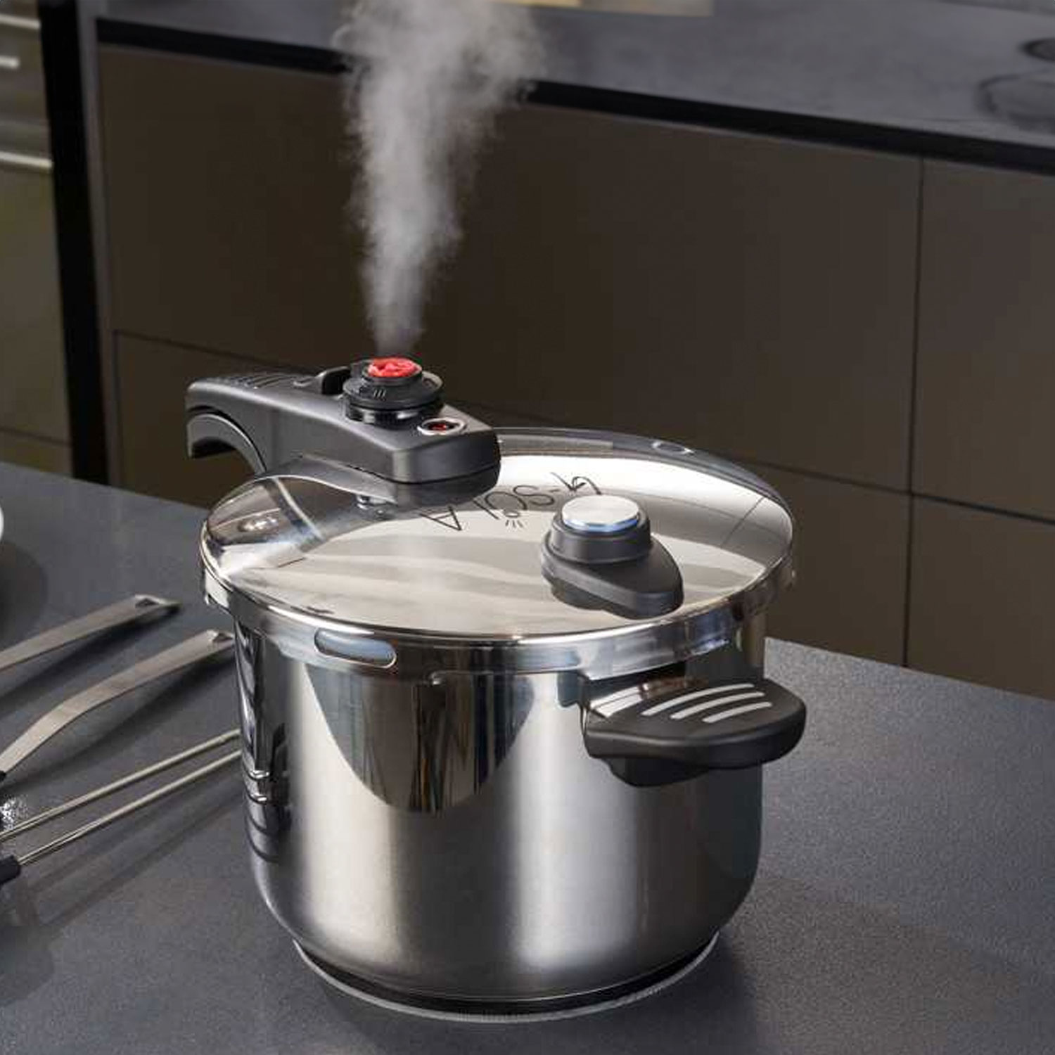 Olla a presión de acero inoxidable con vapor ascendente. Mango negro y pomo rojo. Ambiente de cocina con utensilios.
