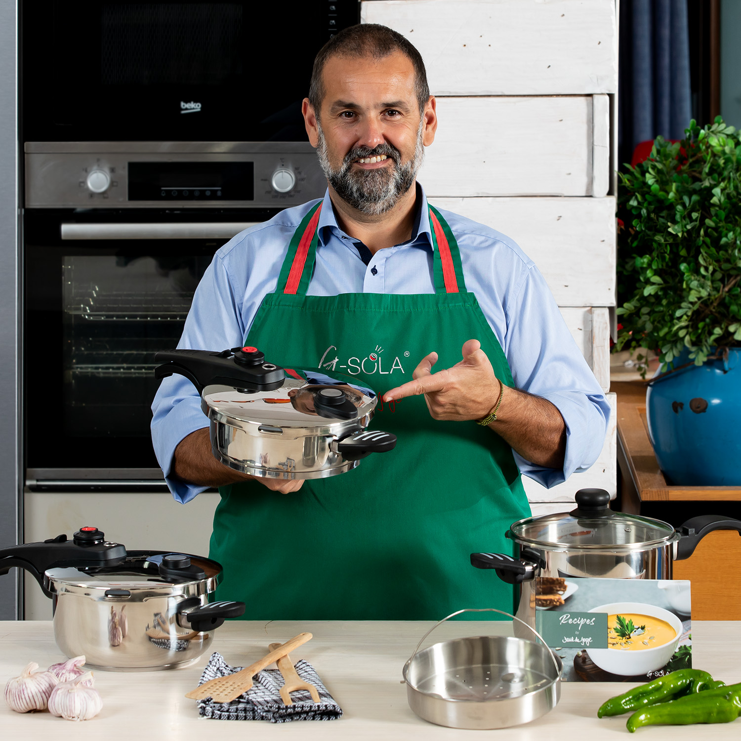 Un hombre con delantal verde señala una olla a presión. Hay más ollas, ajo y recetas sobre la mesa. Un tiesto azul en el fondo.