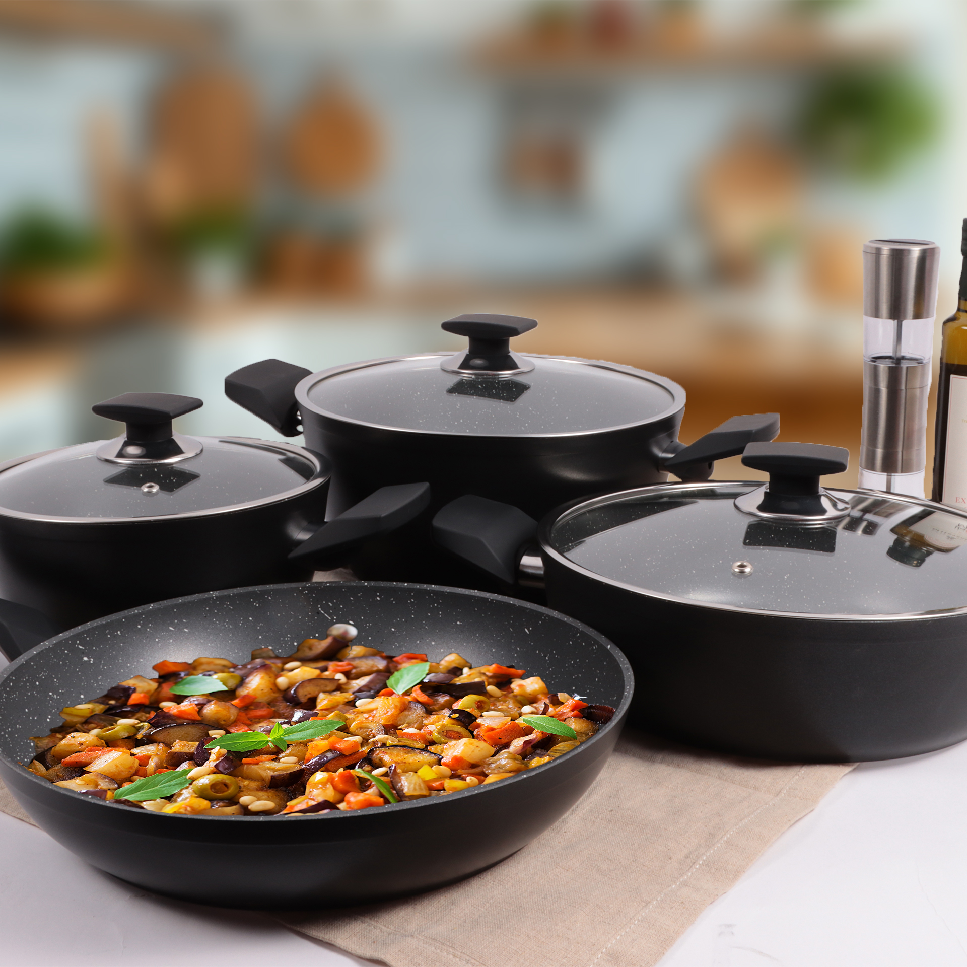 Una sartén con verduras cocidas, rodeada de ollas y sartenes, aceite de oliva y utensilios de cocina sobre una superficie blanca.