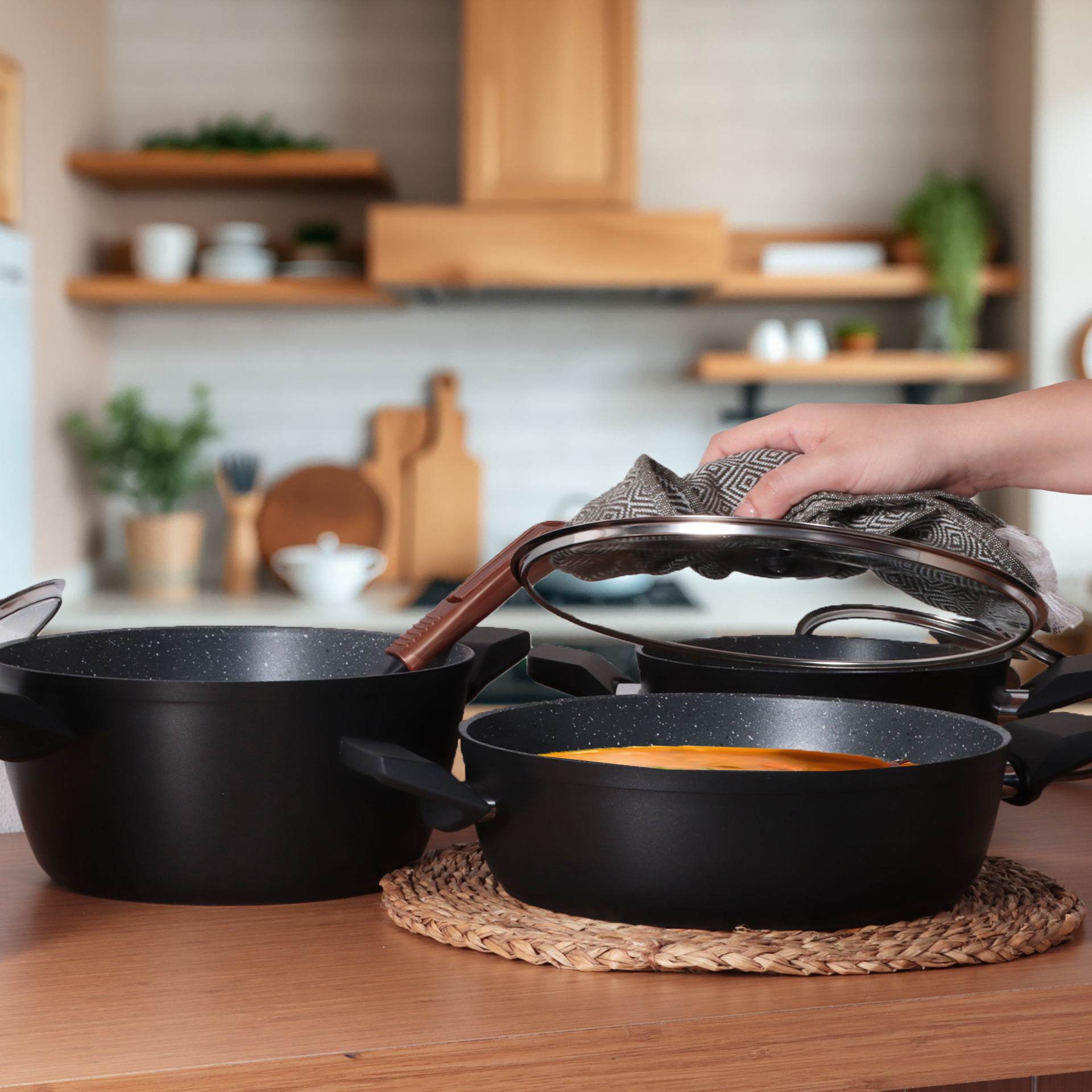 Una mano levanta la tapa de una olla con un paño. Ollas negras sobre una mesa de madera. Cocina al fondo.