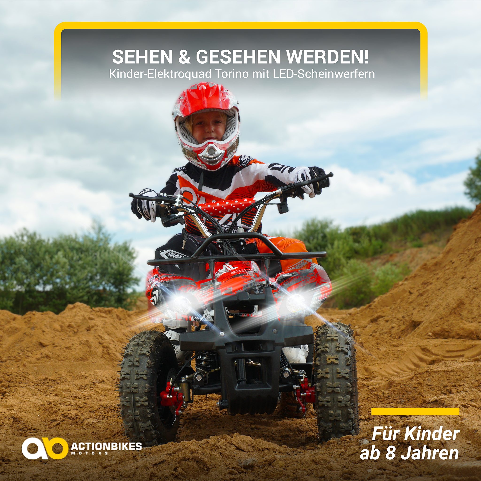 Ein Kind mit rotem Helm fährt ein rotes Quad auf einer sandigen Strecke. Das Quad hat Scheinwerfer. Der Himmel ist sichtbar.