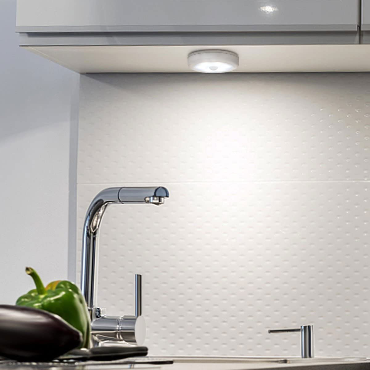 Küchenszene mit Wasserhahn, Gemüse und einer Leuchte unter einem Schrank. Weiße, strukturierte Wand im Hintergrund.
